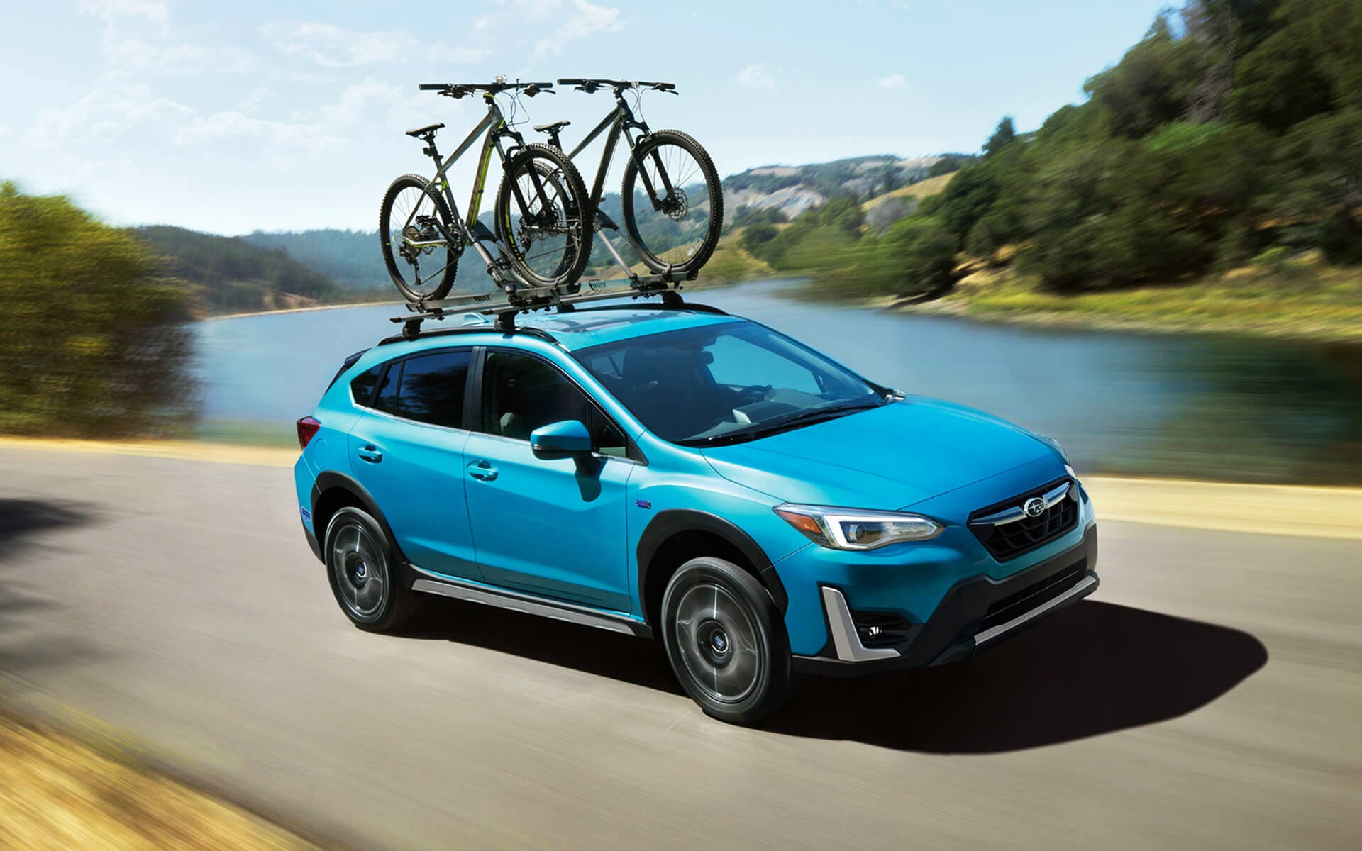 A blue Crosstrek Hybrid with two bicycles on its roof rack driving beside a river | Wyatt Johnson Subaru in Clarksville TN