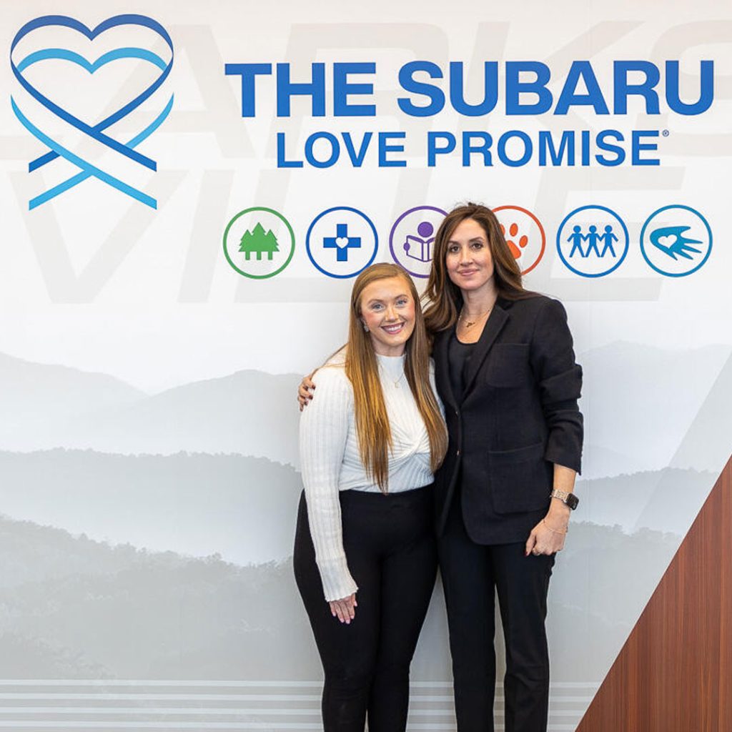 Two women pose in front of a poster for Subaru Love Promise in congruence with Wyatt Johnson Subaru's shoe drive with Operation Warm.